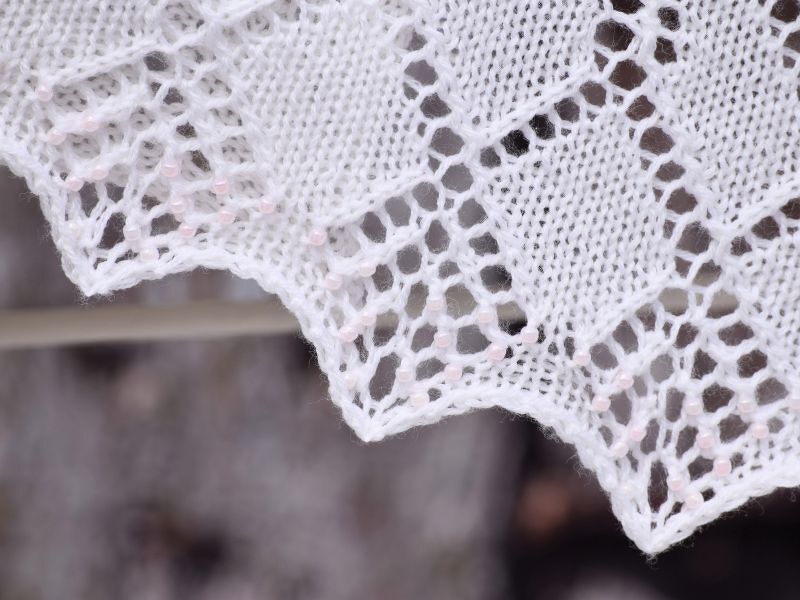 Nahaufnahme weißer Strickkante mit Lochmuster und zartrosa Perlen