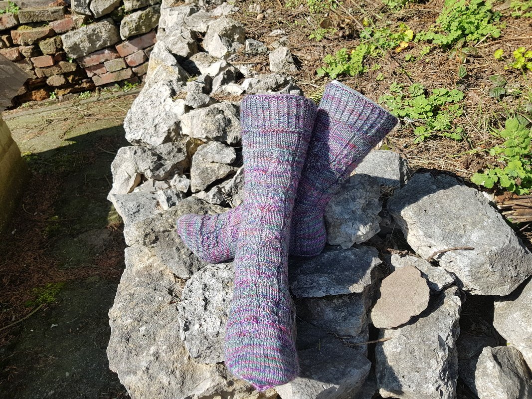Mehrfarbige Stricksocken mit Strukturmuster auf einer Steinfläche vor Ziegeln und Vegetation.