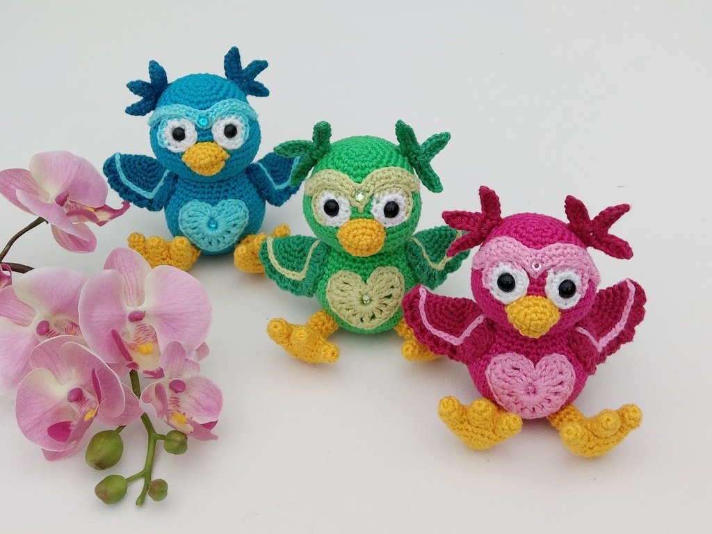 Drei gehäkelte Amigurumi-Eulen in Blau, Grün und Pink mit Herzmotiv am Bauch, großen Augen sowie gelbem Schnabel und gelben Füßen, neben rosafarbenen Orchideen