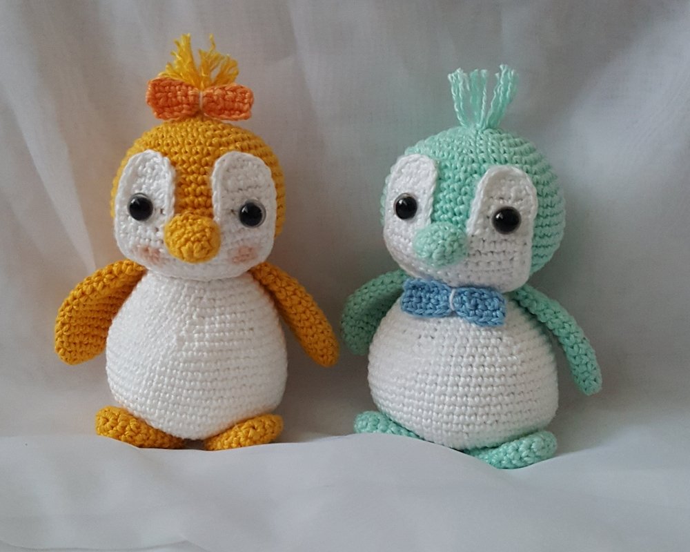 Zwei gehäkelte Pinguin-Amigurumi, einer gelb und einer mint, mit weißem Bauch und schwarzen Augen
