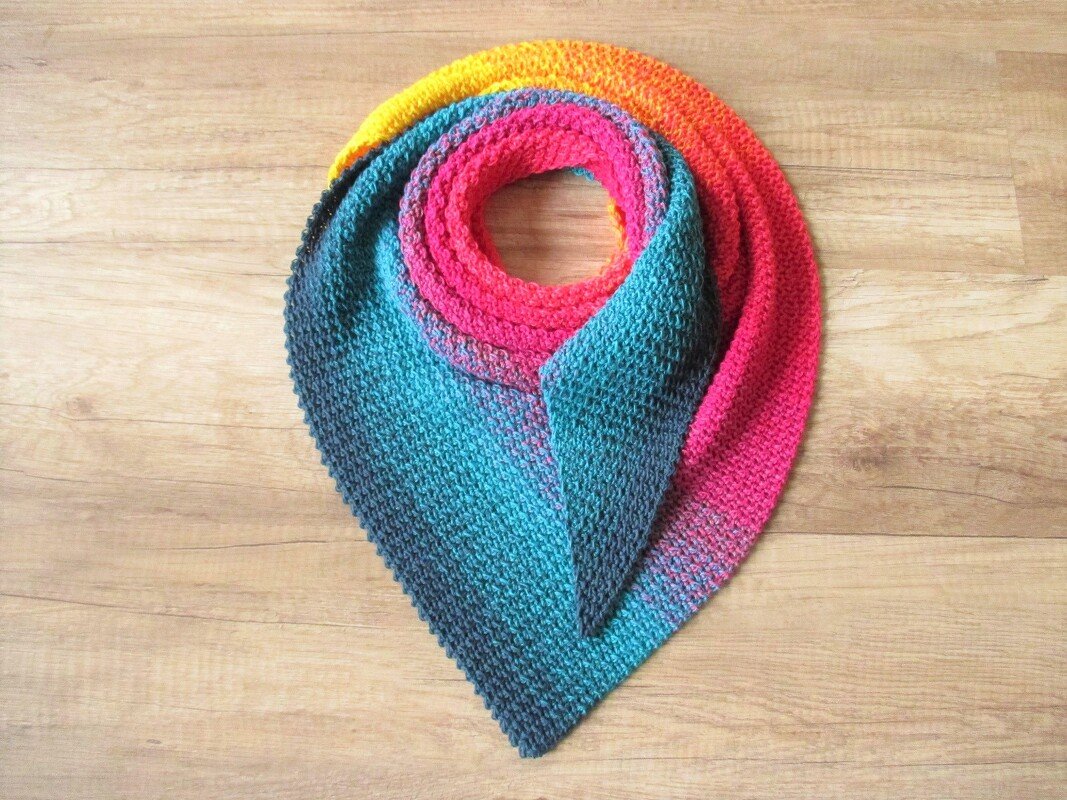 Aufgerolltes gehäkeltes Dreieckstuch mit Farbverlauf von Gelb bis Blau auf Holzuntergrund. Die Maschenstruktur ist gut sichtbar.