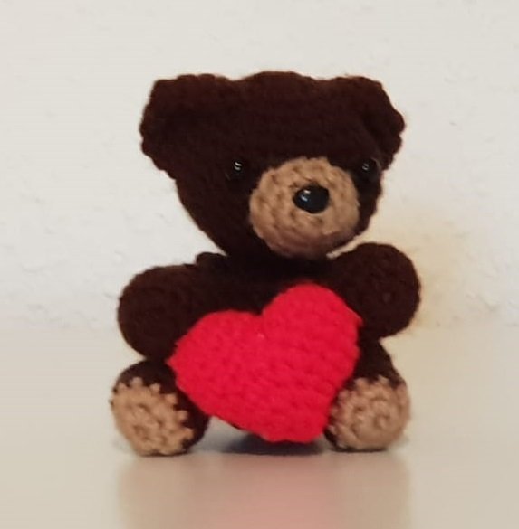 Dunkelbrauner gehäkelter Teddybär mit hellbrauner Schnauze und kleinem roten Herz in den Pfoten.