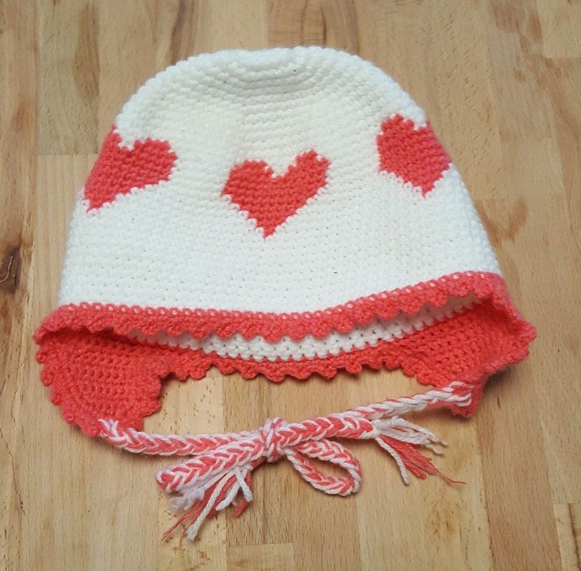 Weiße gehäkelte Babymütze mit korallenfarbenen Herzen, Ohrenklappen und geflochtenen Bindebändern auf Holzuntergrund