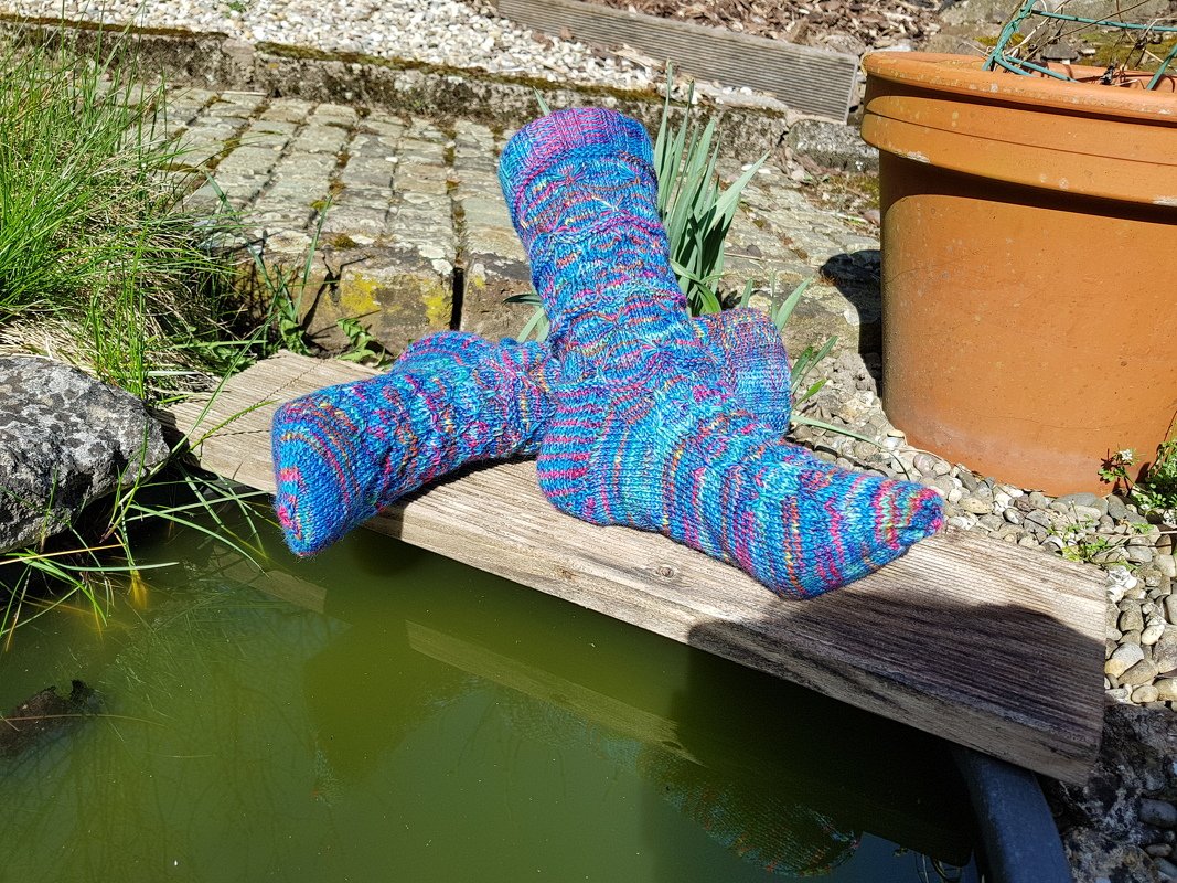Paar handgestrickter Damensocken mit mehrfarbigem Streifenmuster in Blau, Pink und Violett auf einem Holzbrett über einem Teich.