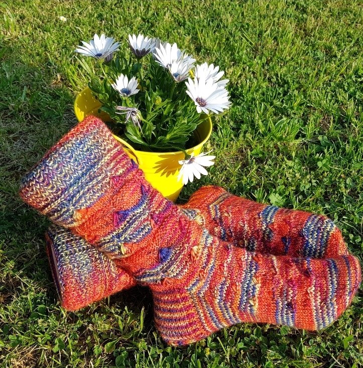 Mehrfarbige gestrickte Damensocken in Orange-Rot auf Gras neben gelbem Blumentopf mit weißen Blumen
