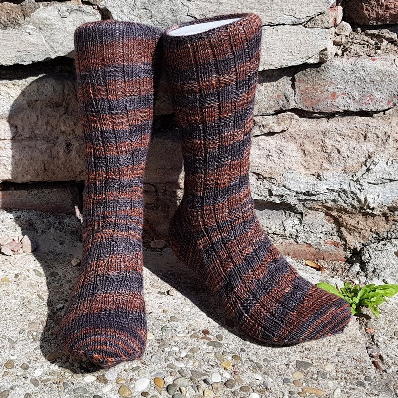 Gestrickte Herrensocken in Braun- und Grautönen mit Streifen lehnen an einer Steinmauer.