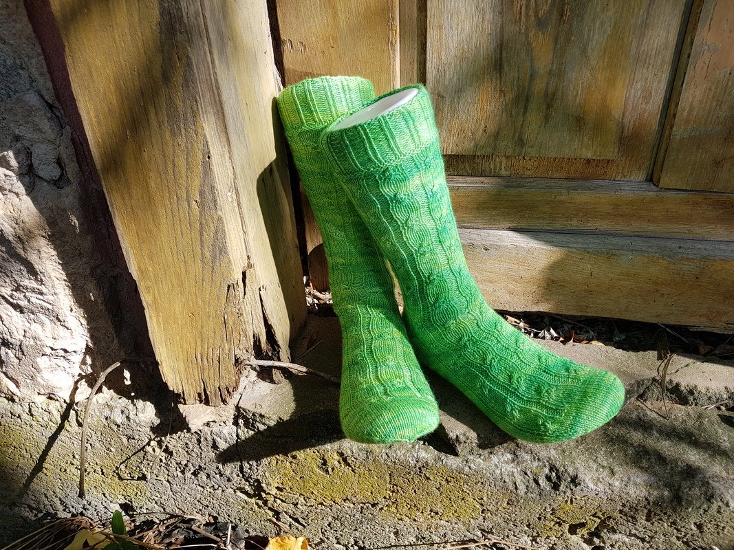 Zwei lange grüne, gestrickte Herrensocken mit Zopfmuster, an eine Holztür gelehnt