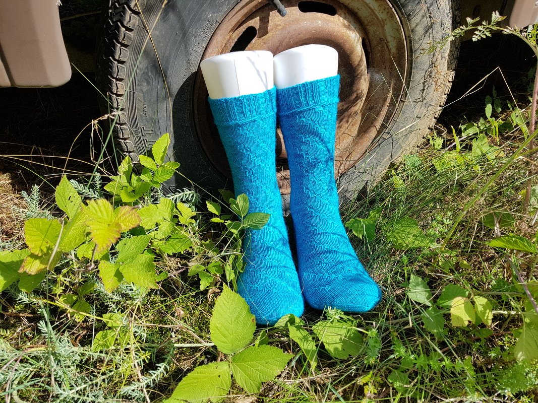 Türkisfarbene gestrickte Herrensocken auf weißen Sockenformen im Gras vor einer rostigen Felge
