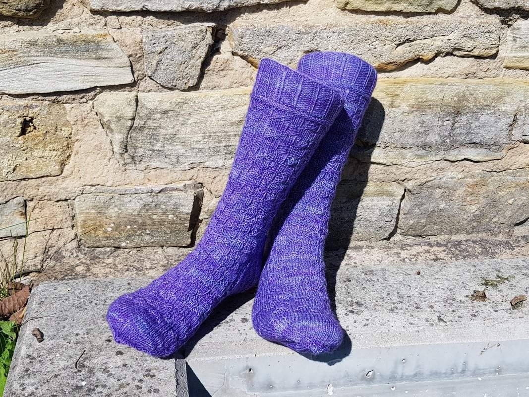Zwei knielange, lila gestrickte Herrensocken mit Strukturmuster vor einer Steinmauer