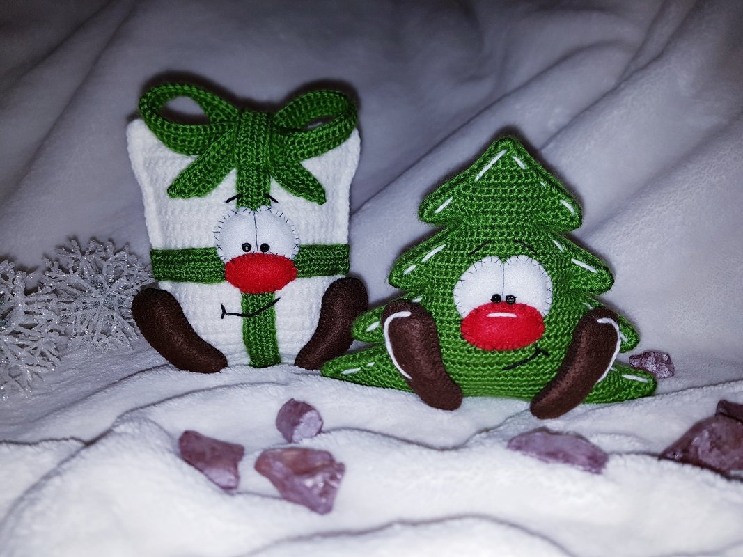 Gehäkeltes Amigurumi-Geschenk mit grünem Band und gehäkelter Tannenbaum mit Gesicht auf weißem Stoff.
