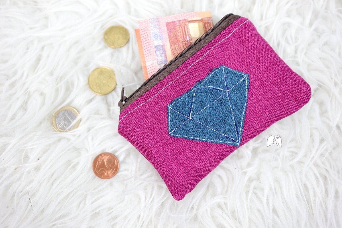 Nahaufnahme einer pinken Stofftasche mit braunem Reißverschluss und blau gestickter Diamant-Applikation, daneben Euro-Münzen und -Scheine auf weißem Untergrund