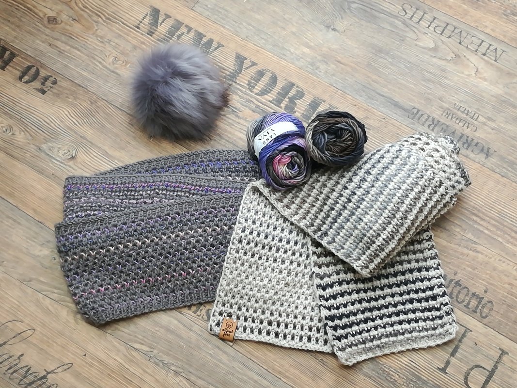 Gestrickte Schals und Loops, zwei Garnknäuel und grauer Bommel auf Holzboden.