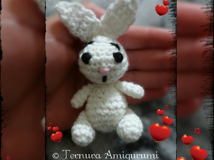 Small white crocheted amigurumi rabbit with black embroidered eyes and pink nose, held in a hand with decorative red heart graphics.