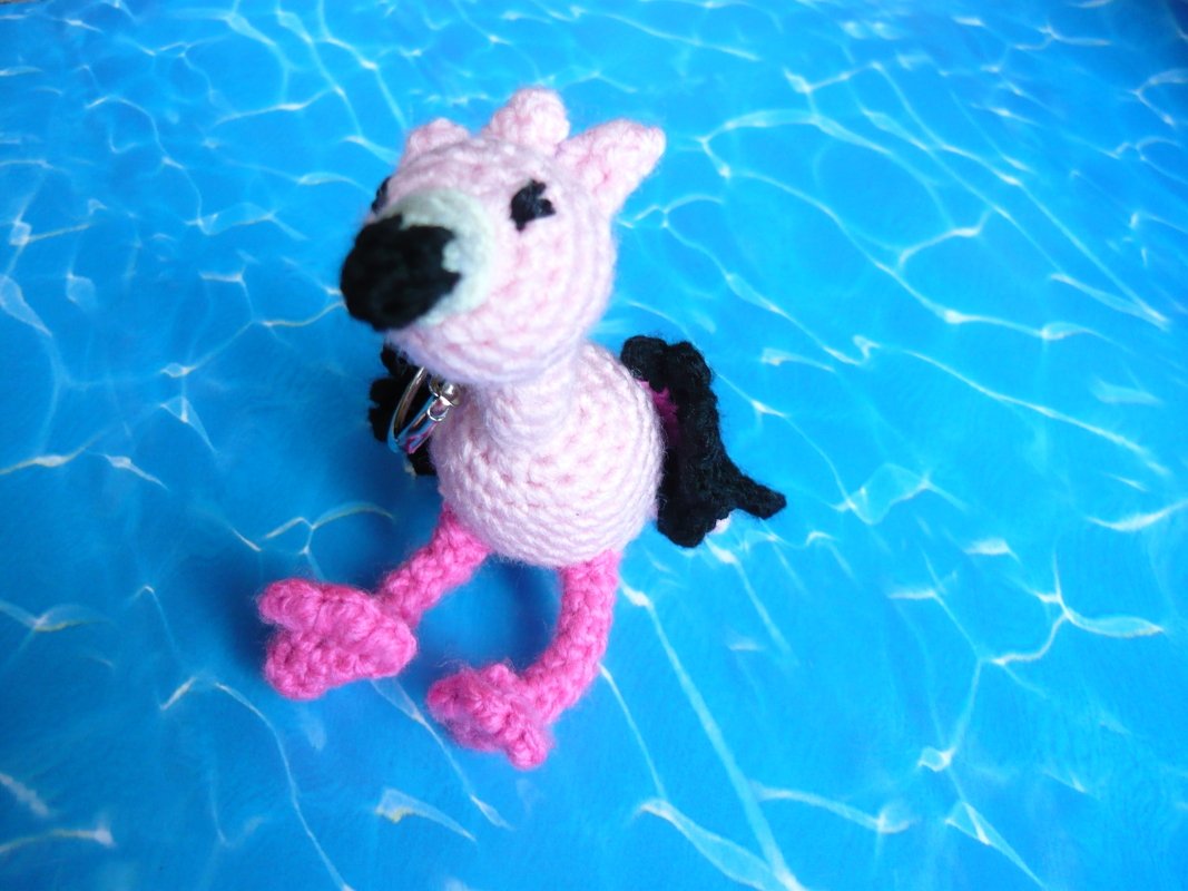 Rosa gehäkelter Flamingo-Amigurumi mit schwarzem Schnabel, schwarzen Flügeln und Metallring vor blauem Hintergrund