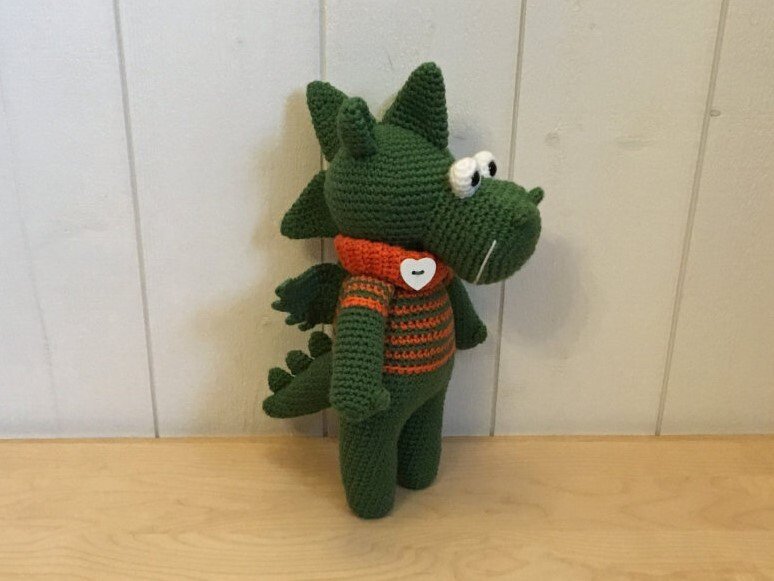 Grüner gehäkelter Amigurumi-Drache mit orange-gestreiftem Schal und Herzknopf, seitliche Ansicht