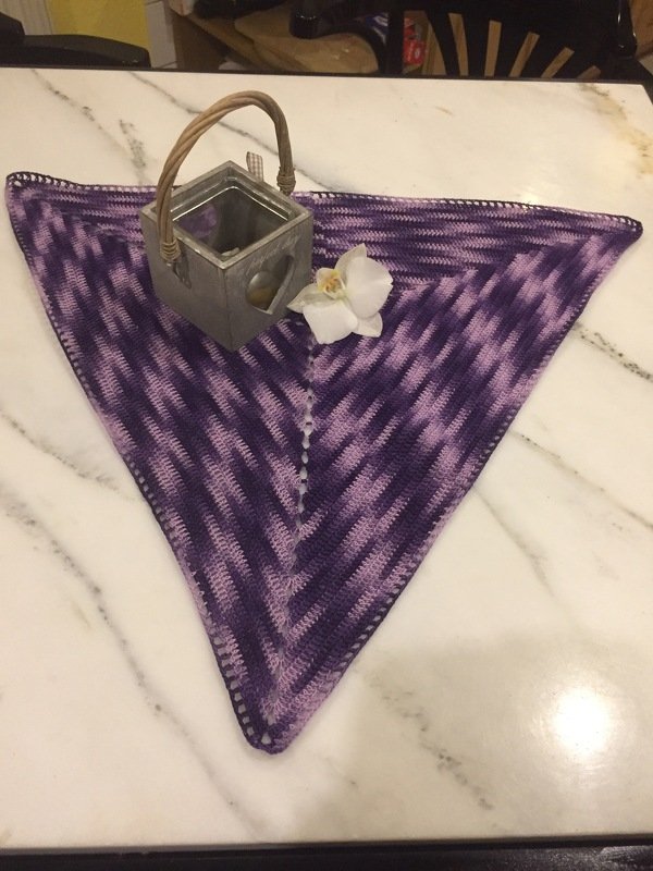 Dreieckiges gehäkeltes Deckchen in Lila- und Violetttönen mit Zickzackmuster auf Marmortisch, dekoriert mit Laterne und weißer Blüte.