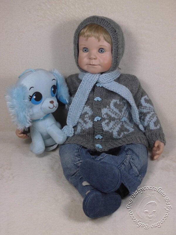 Babypuppe mit grauer Jacke mit hellblauem Muster, passender Mütze und Schal, hält blaues Kuscheltier