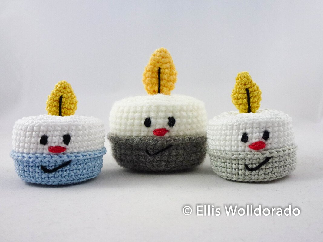 Drei runde gehäkelte Teelicht-Figuren mit Gesichtern und gelben Flammen in Weiß, Hellblau, Dunkelgrau und Hellgrau vor weißem Hintergrund.