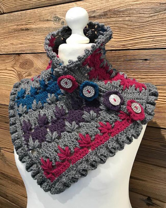 Gehäkelter grauer Kragenschal mit sternförmigen Maschen, pinken, lila und blauen Einsätzen und fünf Zierknöpfen auf einer Büste vor Holzwand.
