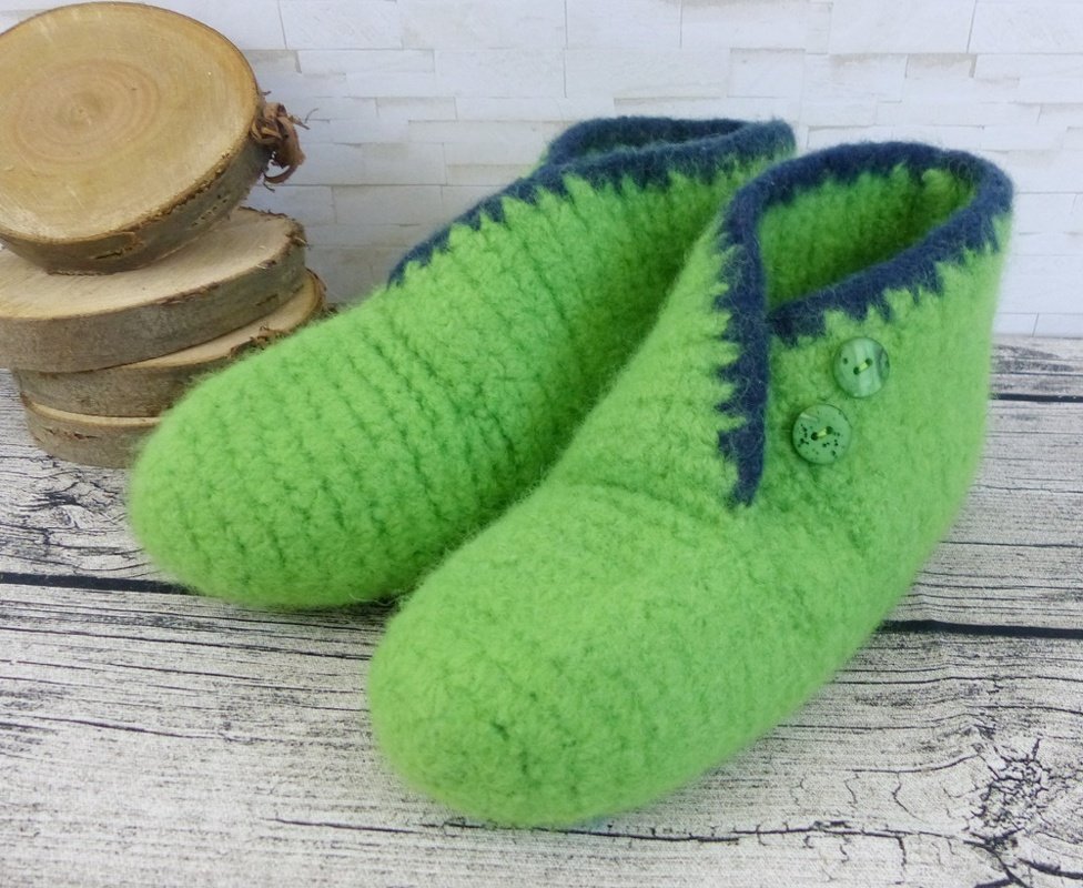 Apfelgrüne, gehäkelte und gefilzte Hausschuhe mit dunkelblauer Borte und zwei Knöpfen auf einem Holztisch.