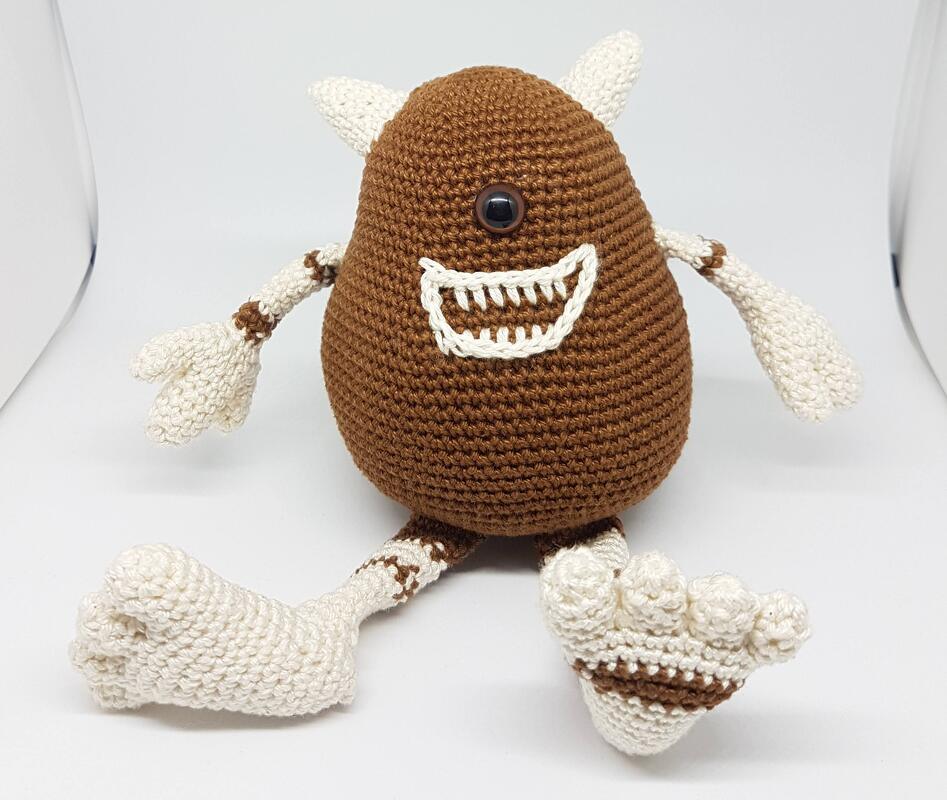Braunes gehäkeltes Amigurumi-Monster mit einem großen Auge, Hörnern, gesticktem Lächeln sowie cremefarbenen Armen und Füßen