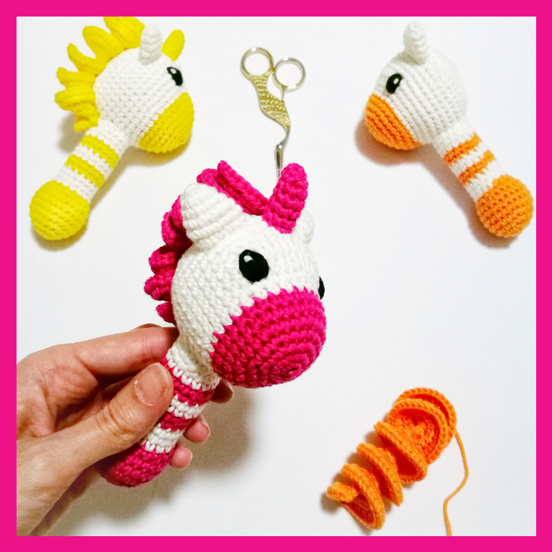 Drei gehäkelte Einhorn-Greiflinge in Pink, Gelb und Orange auf weißem Untergrund; im Vordergrund hält eine Hand ein pink-weißes Exemplar
