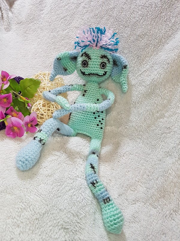 Gehäkeltes mintgrünes und hellblaues Amigurumi-Monster mit Pompom und großen Augen auf hellem Stoff