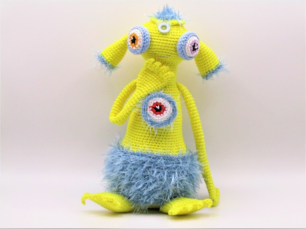 Gehäkeltes gelbes Amigurumi-Monster mit drei großen Augen und blauen flauschigen Akzenten, sitzend