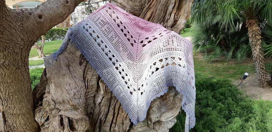 Gehäkeltes Dreieckstuch mit Ombré‑Farbverlauf (Rosa–Weiß–Hellblau) und Lochmuster auf einem Baumstamm.