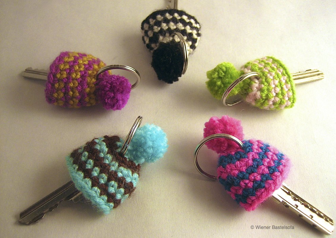Fünf bunte, gehäkelte Schlüsselmützen mit Pompons an Metallschlüsseln in verschiedenen Farbkombinationen.