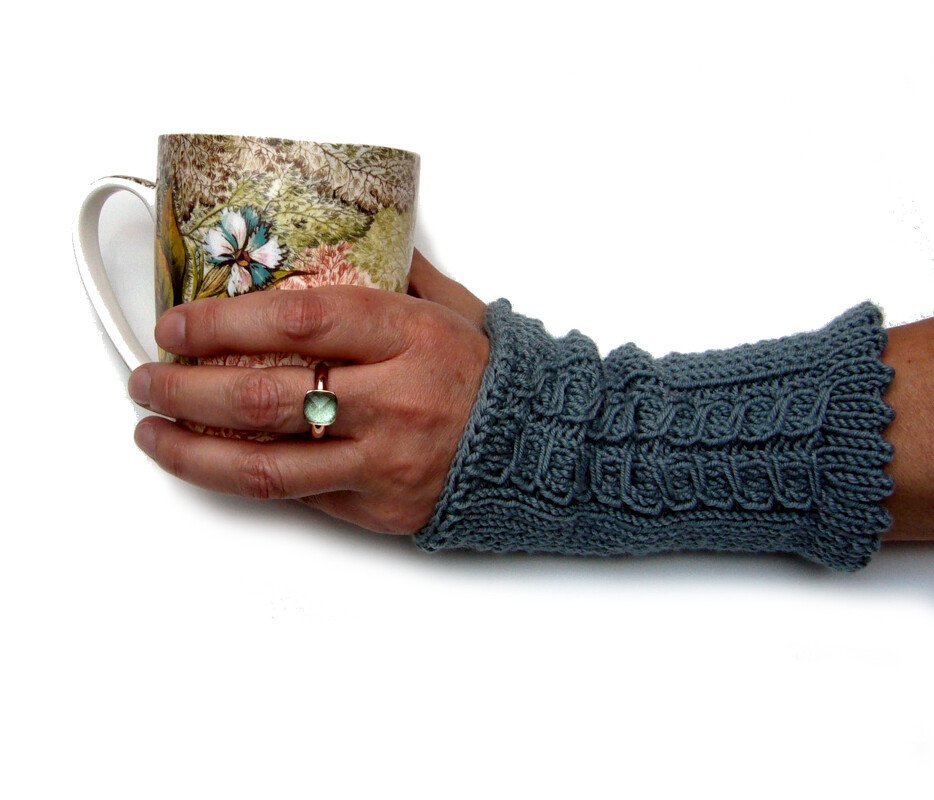 Linke Hand mit graublauen gestrickten Armstulpen mit Zopfmuster, hält eine gemusterte Tasse