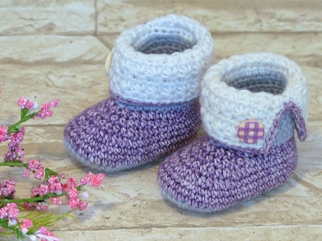 Zwei lavendelfarbene, gehäkelte Babyschuhe mit weißem Umschlag und kariertem Knopf neben kleinen rosa Blüten