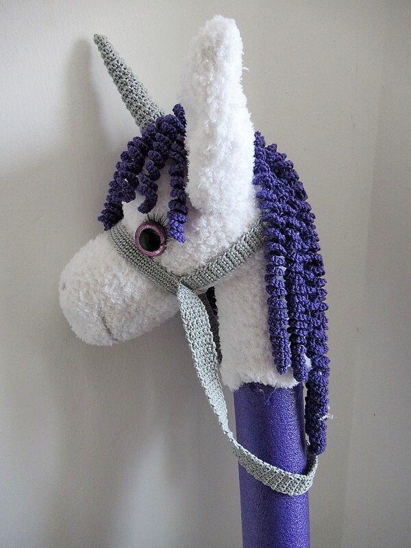 Gehäkeltes weißes Einhorn-Steckenpferd mit lila Mähne, grauem Horn und Zaumzeug auf lila Stab