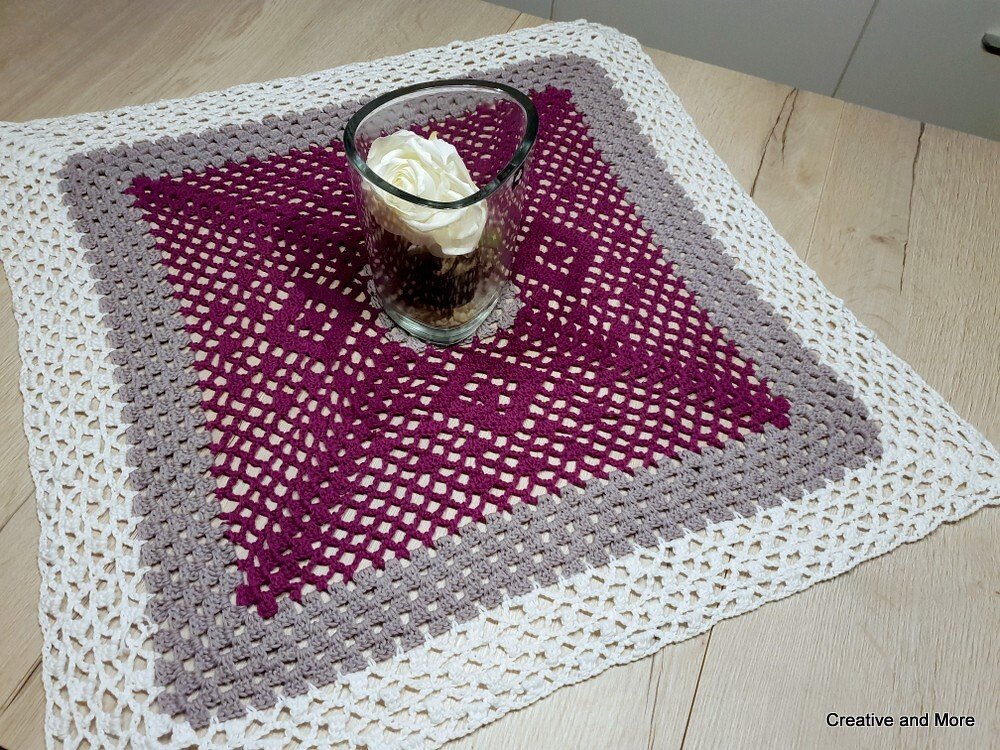 Quadratisches gehäkeltes Deckchen in dunklem Beerenton, Grau und Weiß auf hellem Holztisch mit Glasvase und weißer Rose