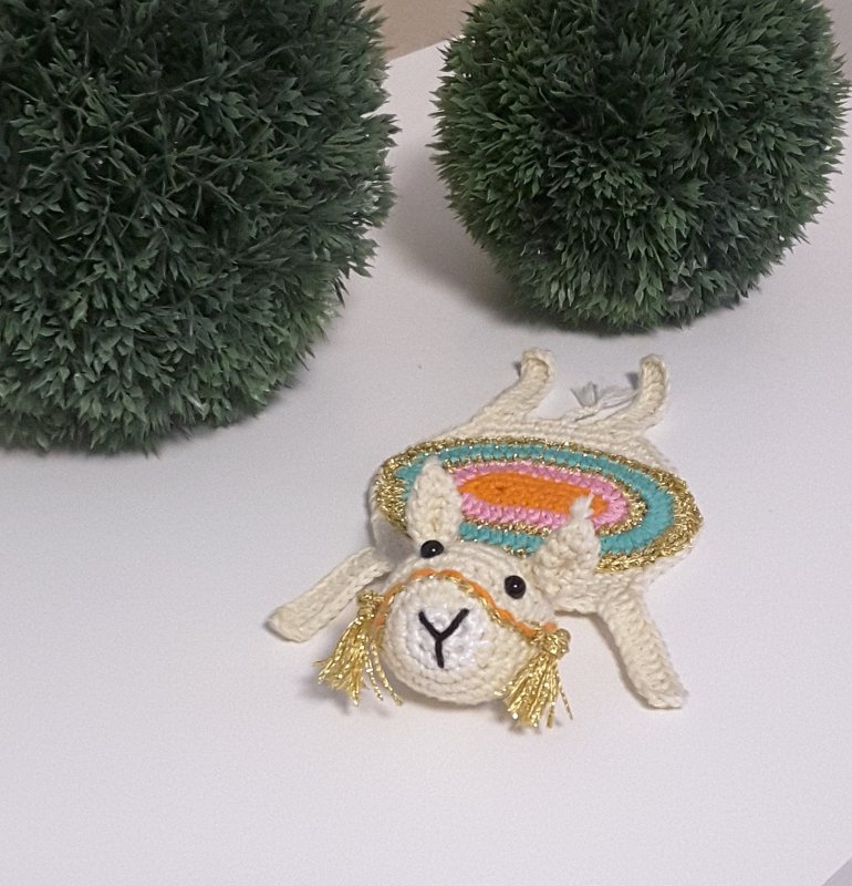 Gehäkelter Lama-Untersetzer in Creme mit türkis-rosa-orangefarbigem Sattel und goldenen Fransen, auf weißer Oberfläche neben zwei grünen Dekokugeln