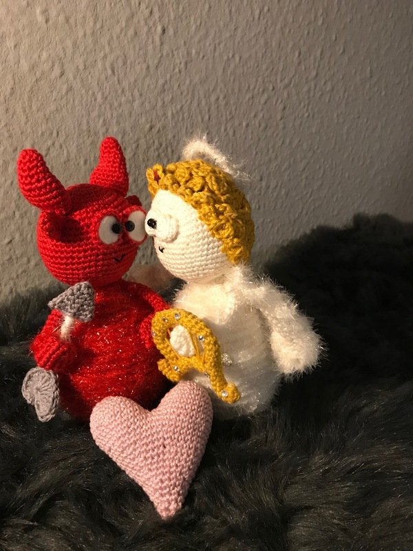 Rotes gehäkeltes Teufelchen und weißes gehäkeltes Engelchen mit rosa Herz auf dunklem Plüsch.
