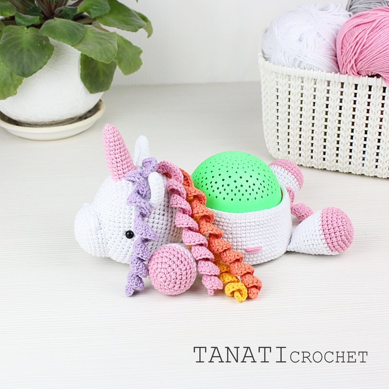 weißes gehäkeltes Einhorn liegend mit rosa Horn, mehrfarbiger Spiralmähne und grünem Kuppel-Einsatz
