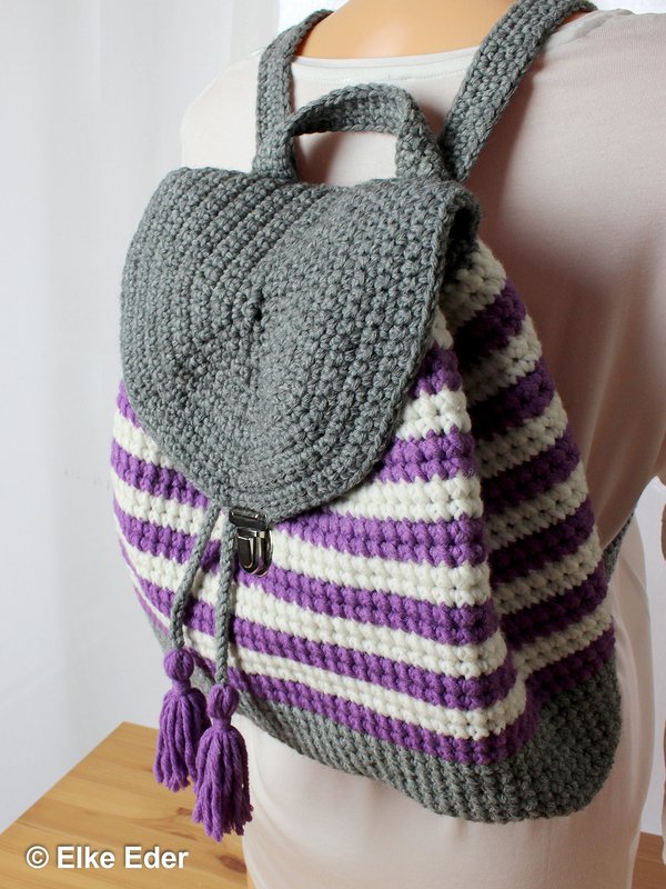 Back view of a person wearing a crocheted backpack with a grey flap and purple and white stripes.