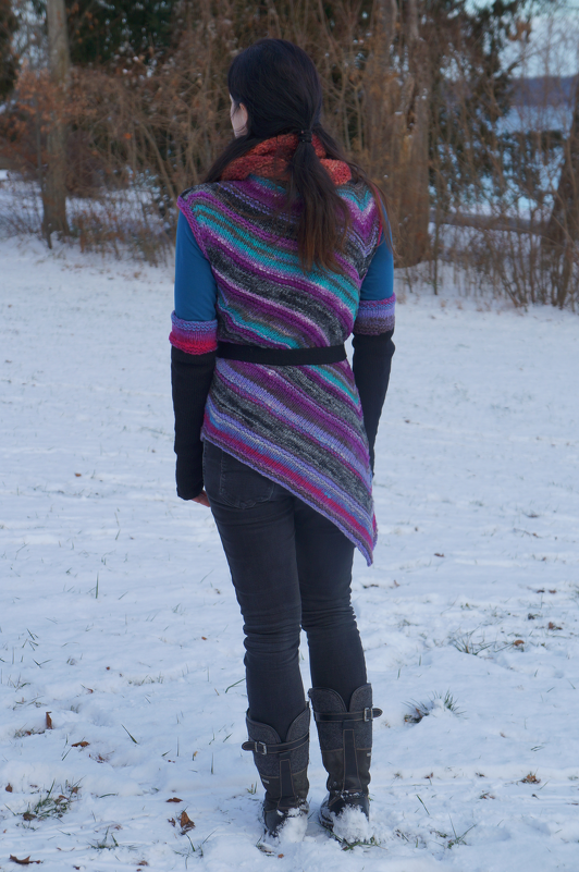 Rückansicht einer Person im Schnee mit mehrfarbigem gestrickten Wickelponcho, diagonalen Streifen, Gürtel und asymmetrischem Saum