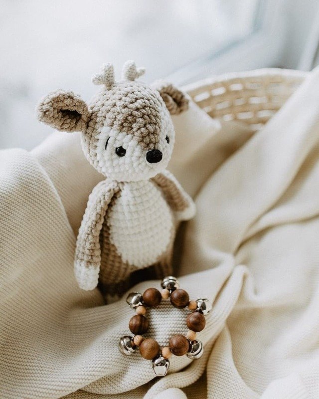Gehäkeltes Amigurumi‑Reh in Beige auf einer Strickdecke neben einer Holzperlenrassel mit Glöckchen.
