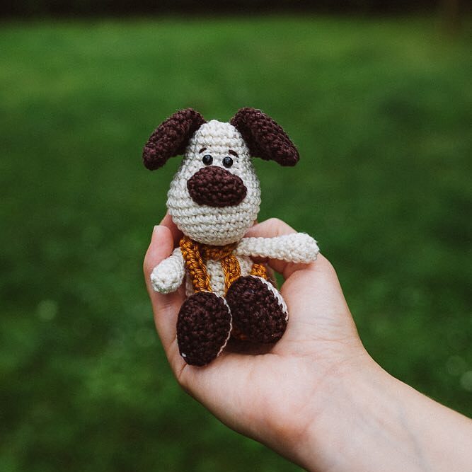Hand hält kleinen beigen und braunen Amigurumi-Hund mit goldfarbenem Schal vor grünem Hintergrund.