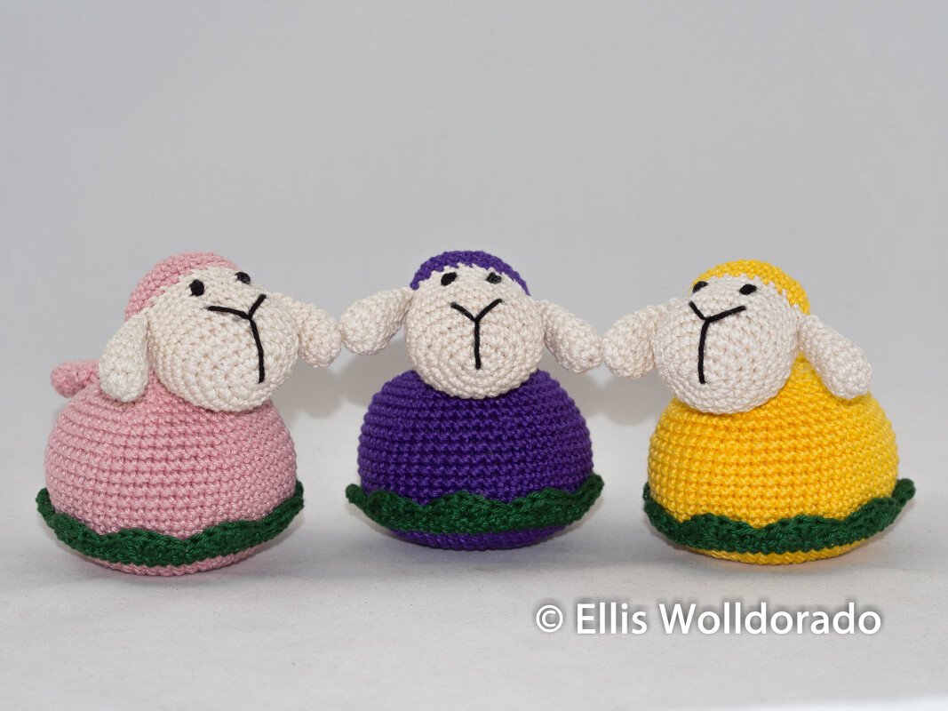 Drei gehäkelte Amigurumi-Lämmer in Rosa, Violett und Gelb nebeneinander auf hellem Hintergrund