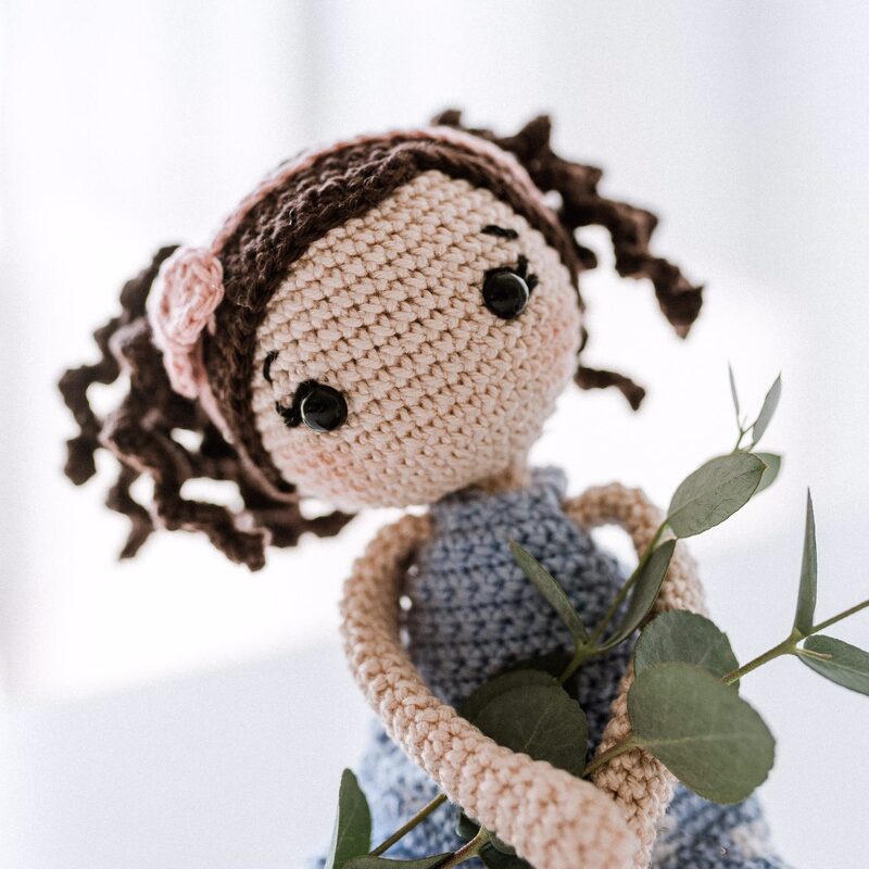 Gehäkelte Amigurumi-Puppe mit braunen Locken, blauem Kleid und Eukalyptuszweig