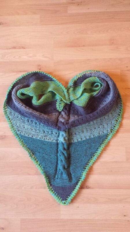 Gestricktes Dreieckstuch in Grün- und Blautönen mit zentralem Zopfmuster, Lochreihen und kontrastfarbiger Einfassung, herzförmig auf Holzboden drapiert.