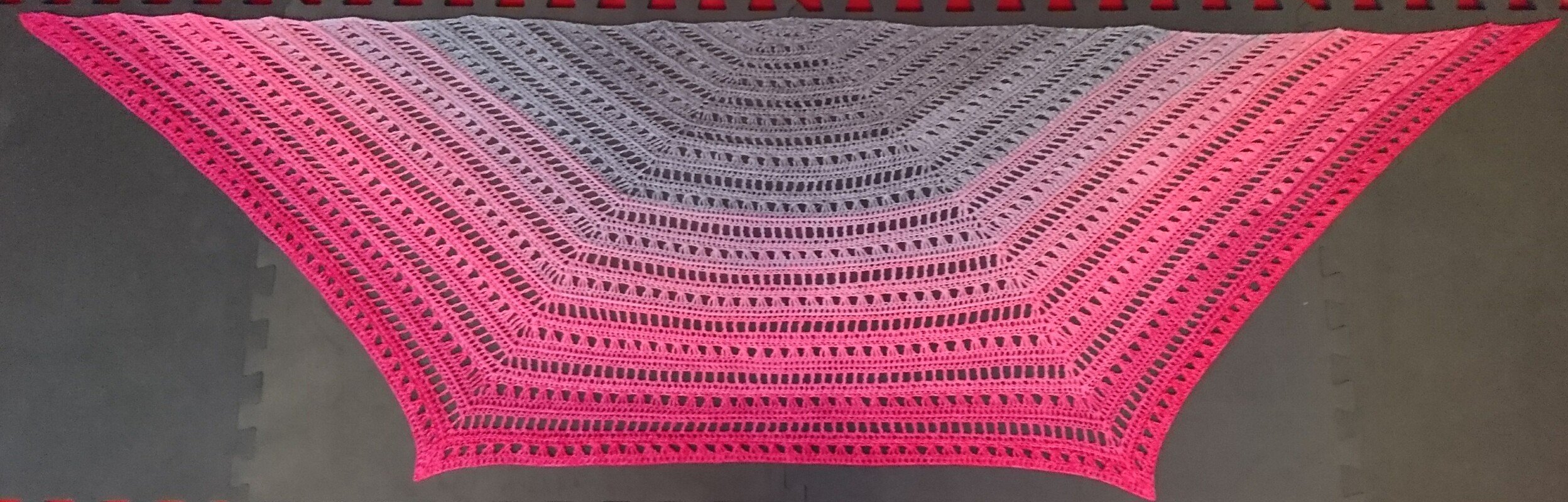 Gehäkeltes halbrundes Tuch mit Farbverlauf von Pink zu Grau, ausgebreitet auf dunklem Untergrund