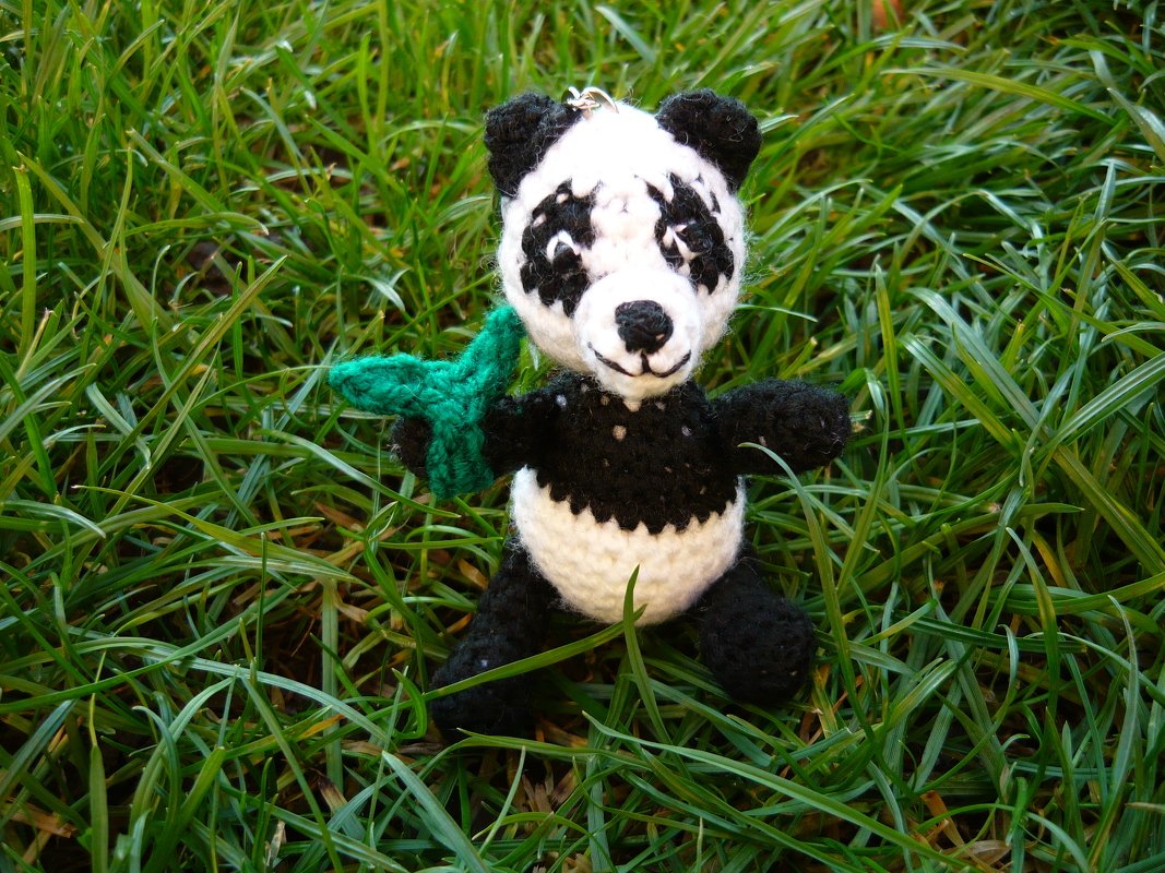 Gehäkelter schwarz-weißer Panda-Amigurumi sitzt im Gras mit kleinem grünen Blatt und Anhängerschlaufe