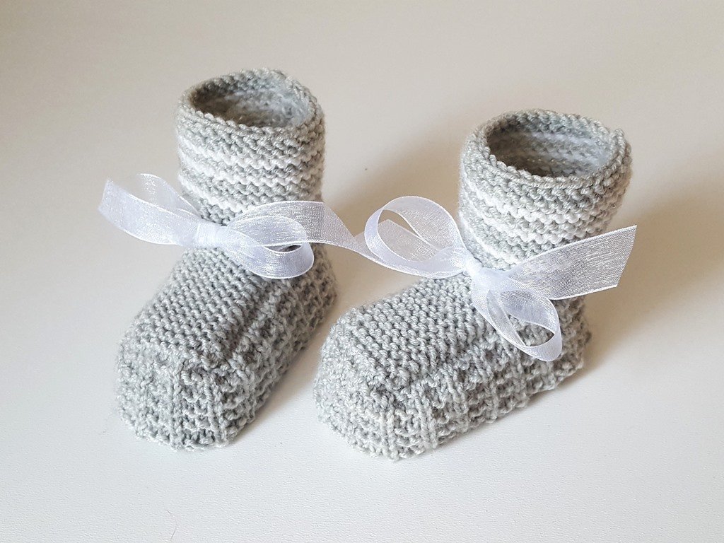 Zwei handgestrickte hellgraue Babyschuhe mit feinen weißen Streifen und weißen Schleifen, Nahaufnahme des Strickmusters und der Verarbeitung.