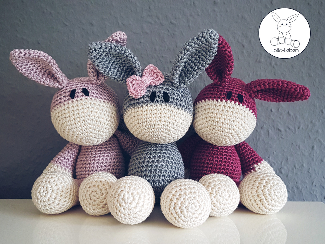 Three crocheted amigurumi bunnies in pink, grey, and burgundy with cream faces and round paws seated side by side; the grey bunny wears a small pink bow.