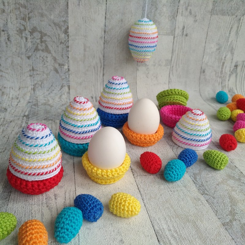 Gehäkelte Osterdekorationen: gestreifte Eierwärmer, Eier in gelben und orangefarbenen Häkelringen und verstreute kleine Amigurumi‑Eier auf hellem Holz