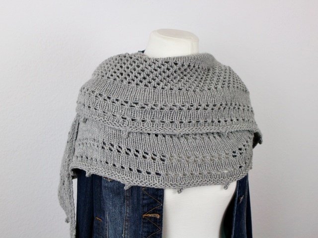 Grey knitted crescent shawl with lace and openwork draped on a mannequin over a denim jacket.